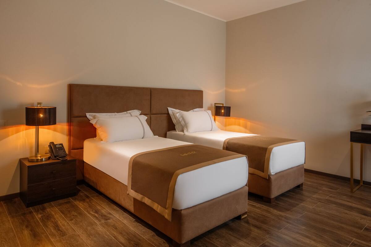 Ein Hotelzimmer mit zwei Betten, jeweils mit brauner Kopfteil und weißer Bettwäsche. Am linken Bett befindet sich ein Nachttisch mit einer Lampe und einem Telefon.