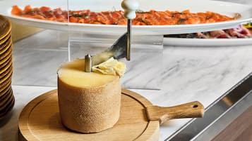 A cheese wheel with a carving tool, a wooden cutting board, and a plate of sliced cheese in the background.
