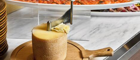 A cheese wheel with a carving tool, a wooden cutting board, and a plate of sliced cheese in the background.