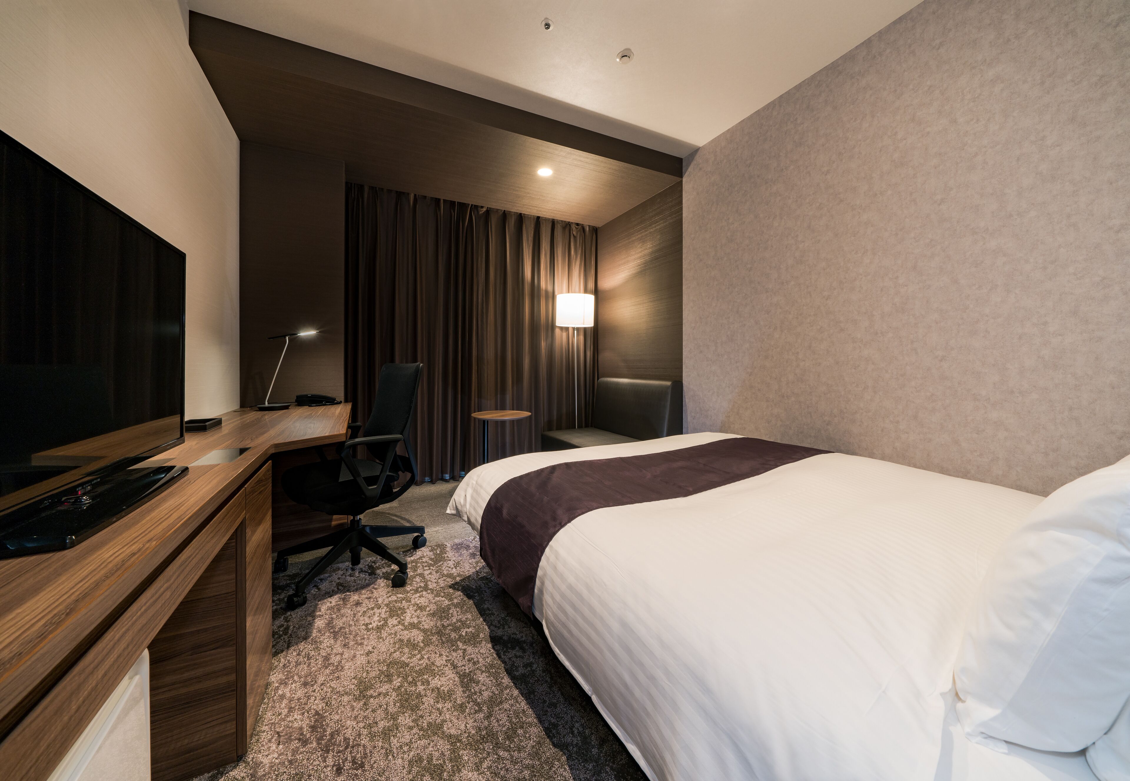 A hotel room with a bed, a desk with a computer, a chair, a TV, and a lamp.
