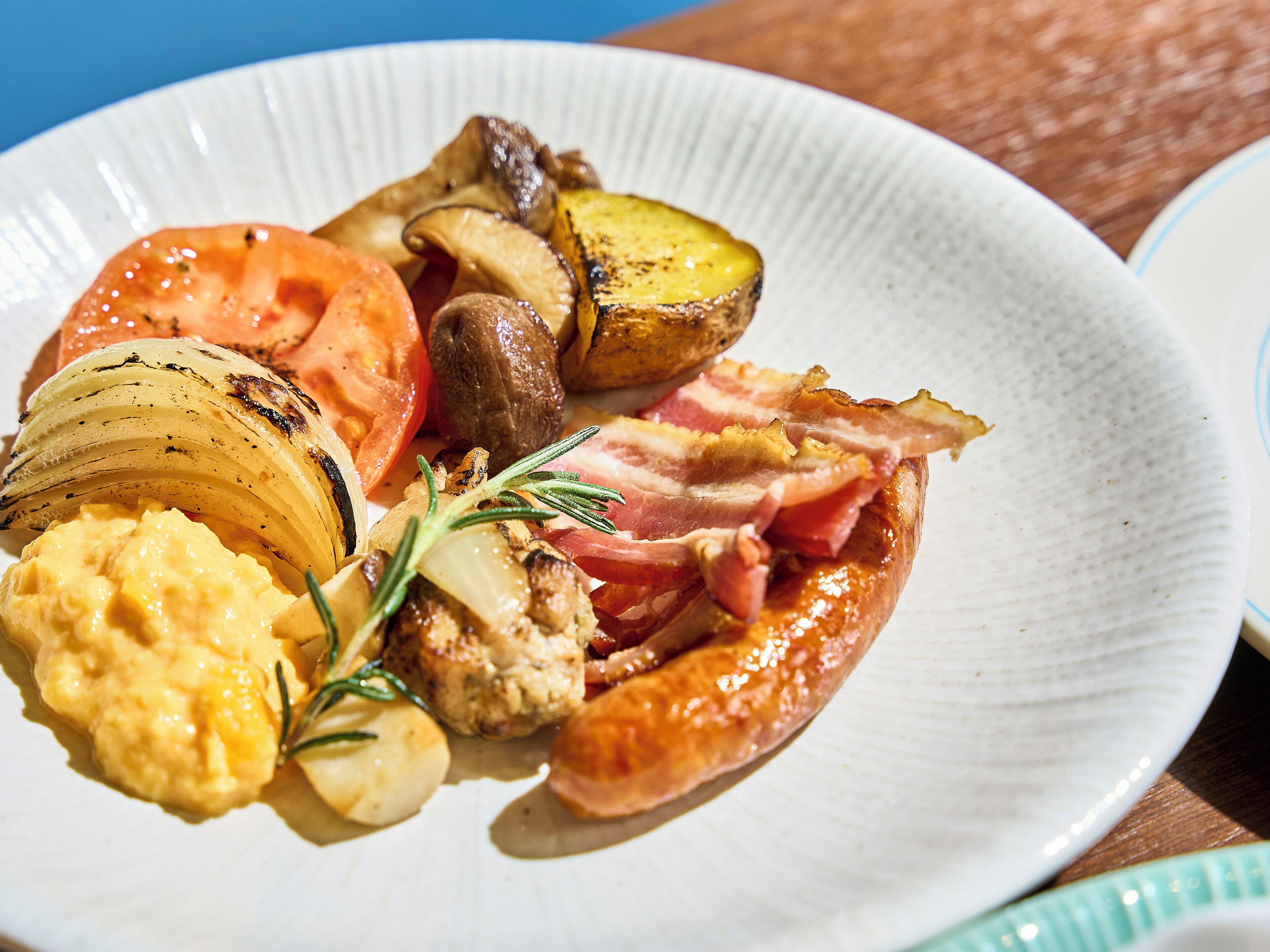 A plate of food with grilled vegetables, bacon, and a sausage.