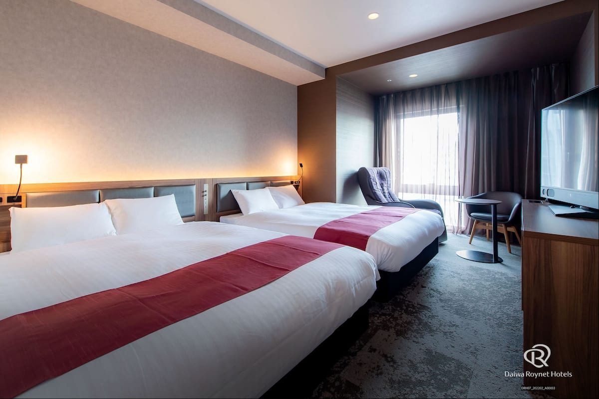 Une chambre d’hôtel avec deux lits, un bureau avec une chaise, une télévision et une fenêtre avec des rideaux.