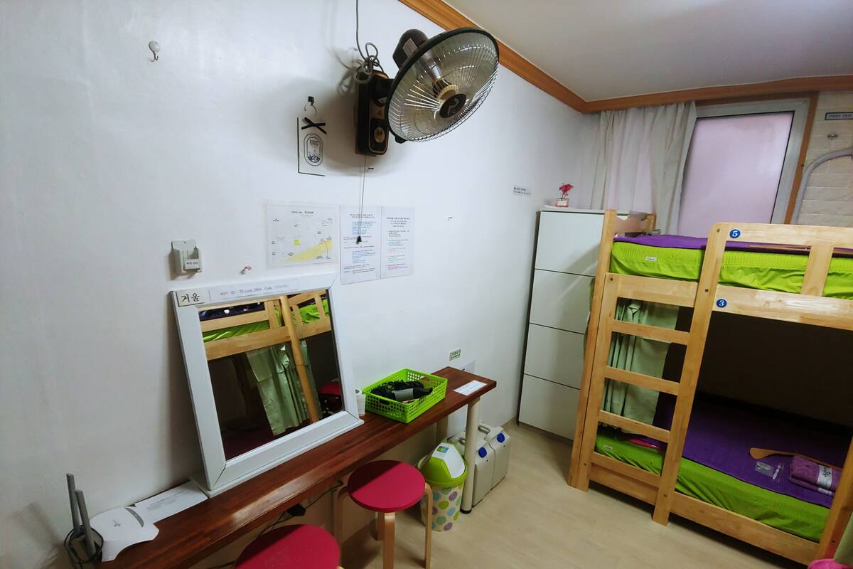 Una habitación con literas, un escritorio con dos taburetes rojos, un ventilador y un refrigerador.