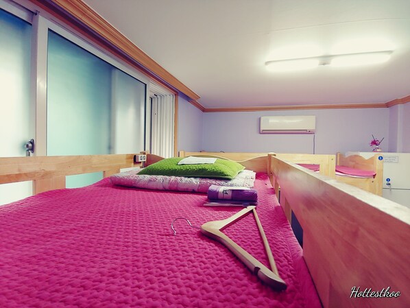 Una habitación con una cama, cabecero de madera, una ventana con persianas, unidad de aire acondicionado y una mesita con un florero.