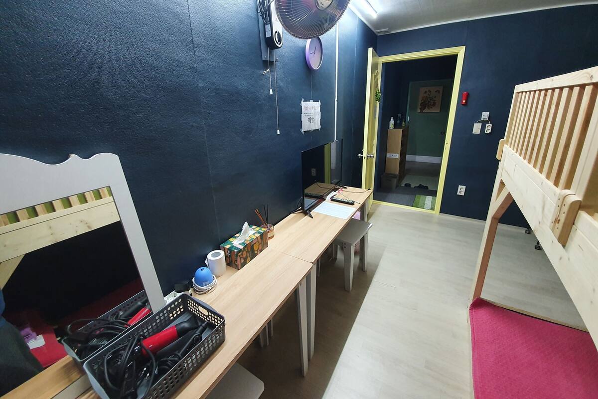 Una habitación con una cama litera, un escritorio con computadora, un ventilador y una puerta que da a otra habitación.