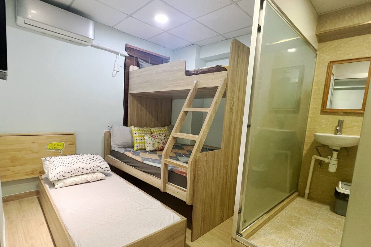 A dormitory room with a bunk bed, a single bed, a small bathroom with a sink, and a shower.