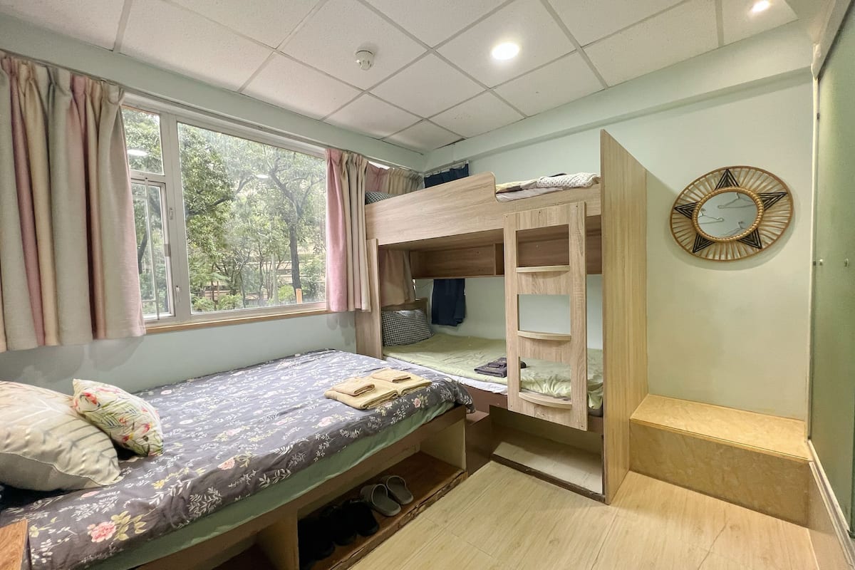 A dormitory room with bunk beds, a window with curtains, a bed with a floral bedspread, a wooden wardrobe, and a wall clock.