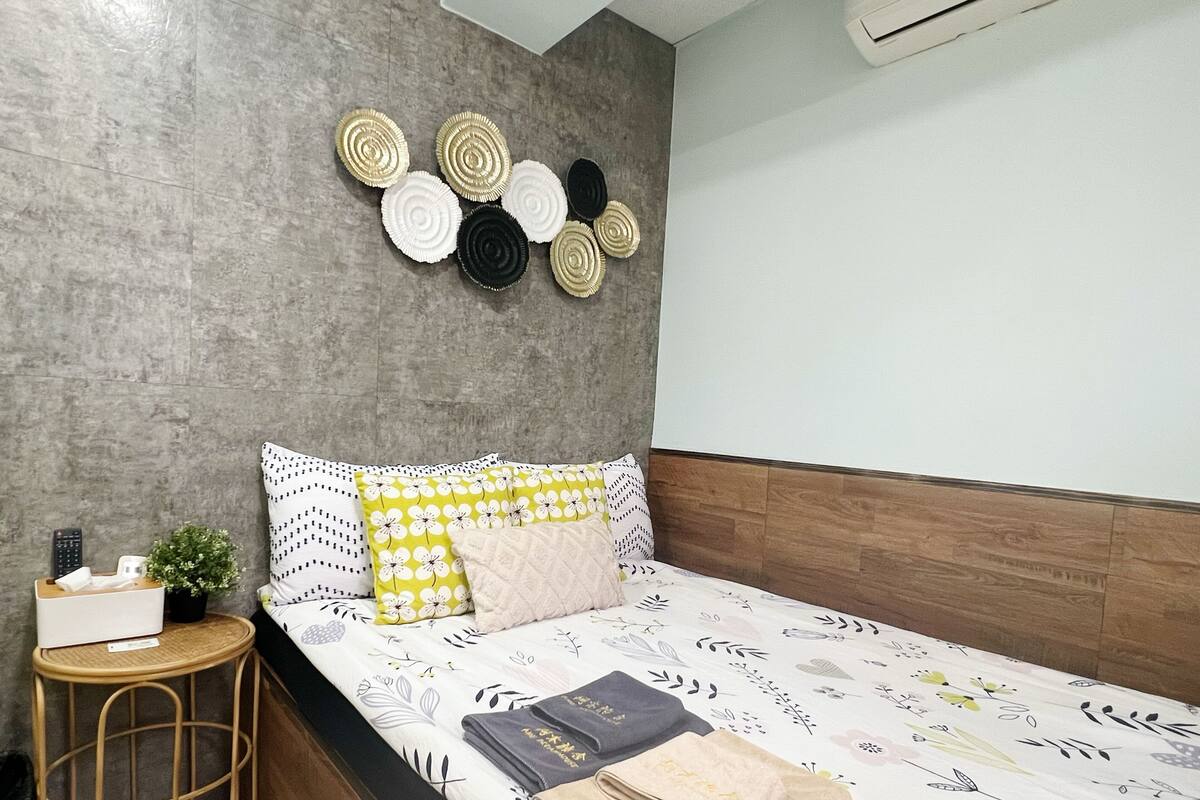 A neatly made bed with floral bedding, a bedside table with a remote control, a small plant, and a wall-mounted decorative item.