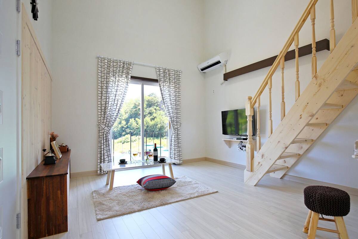 A modern living room with a wooden staircase, a TV mounted on the wall, and a large window with curtains.
