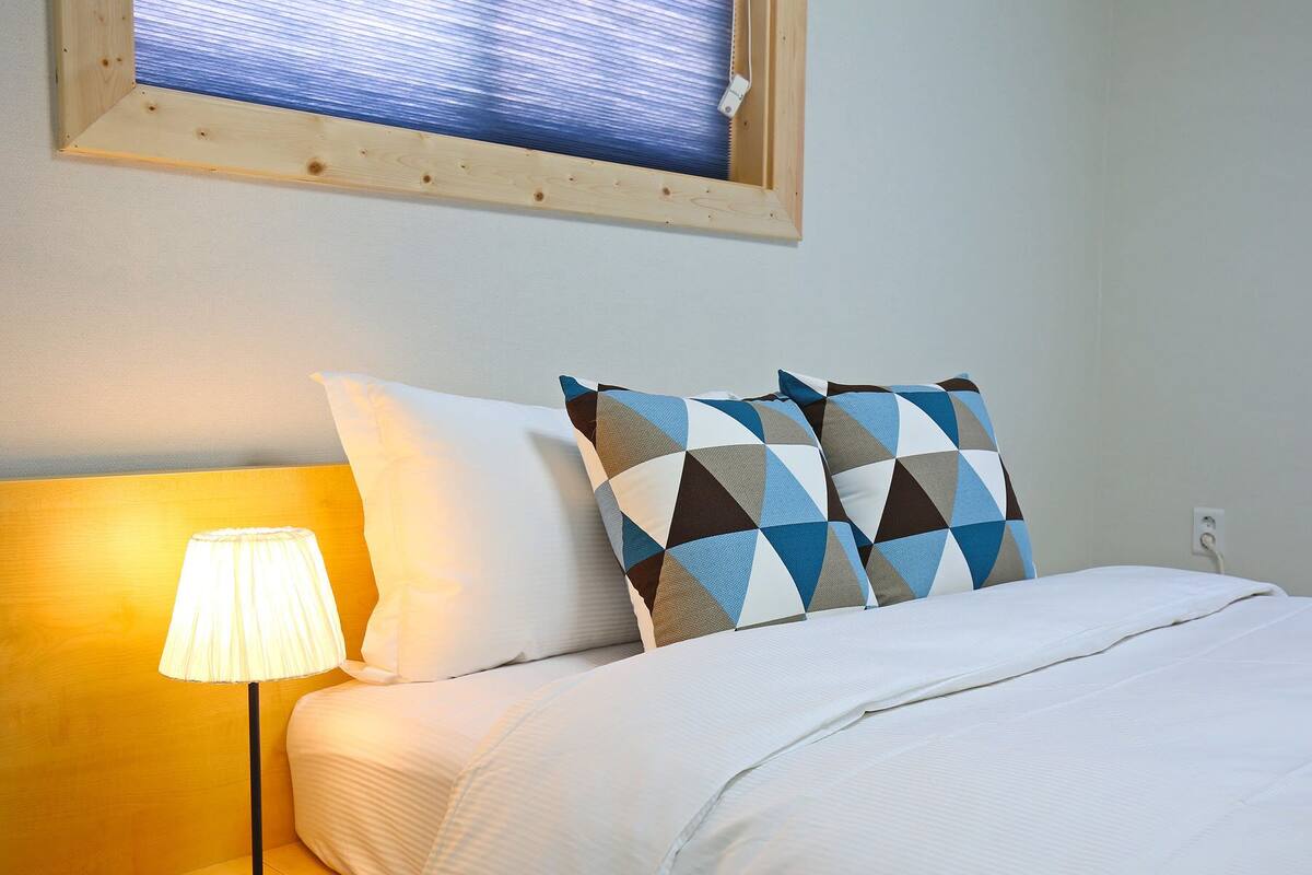 A neatly made bed with white bedding, a lamp on the left, and a window with a view of the sky.