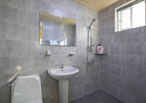 A bathroom with a white sink, a shower, and a toilet. There is a mirror above the sink and a window on the right side.