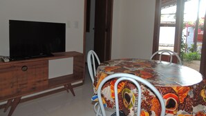 A room with a TV on a wooden cabinet, a round table with a coffee-themed tablecloth, and a door leading outside. - Our House in Jeri 1 (Jijoca de Jericoacoara)