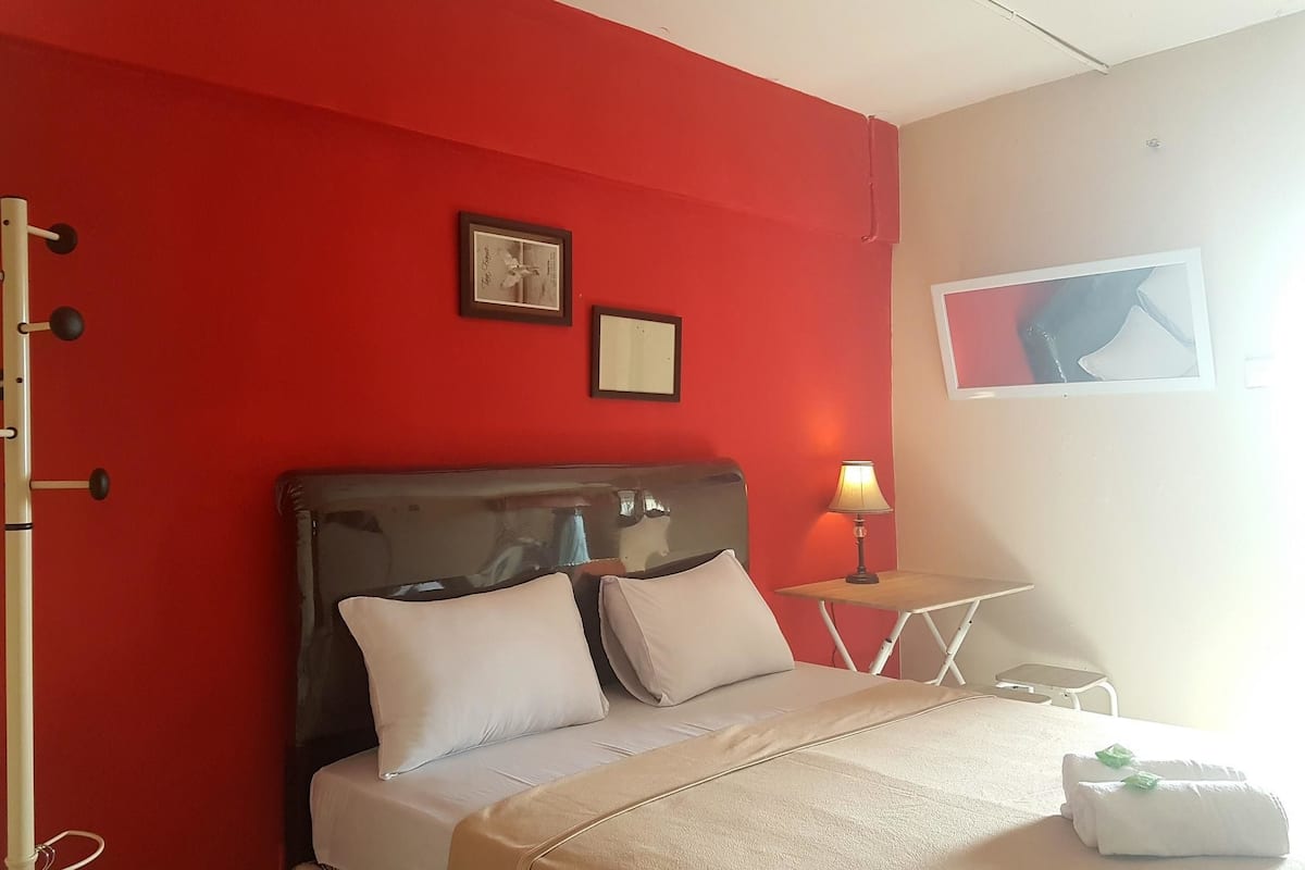 Ein Hotelzimmer mit roter Akzentwand, einem Bett mit weißer Bettwäsche, einem kleinen Tisch mit Lampe und einem gerahmten Bild an der Wand.