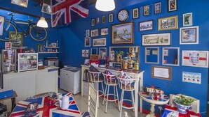 Ein Zimmer mit blauer Wand, verziert mit gerahmten Bildern und einer großen Union Jack Flagge. Weiße Barhocker, ein kleiner Tisch mit Union Jack Tischdecke und ein Fahrrad, das an der Wand hängt.