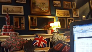 Ein Zimmer mit Schreibtisch, Computer und Stuhl. An der Wand befinden sich gerahmte Bilder und auf dem Schreibtisch eine Lampe.
