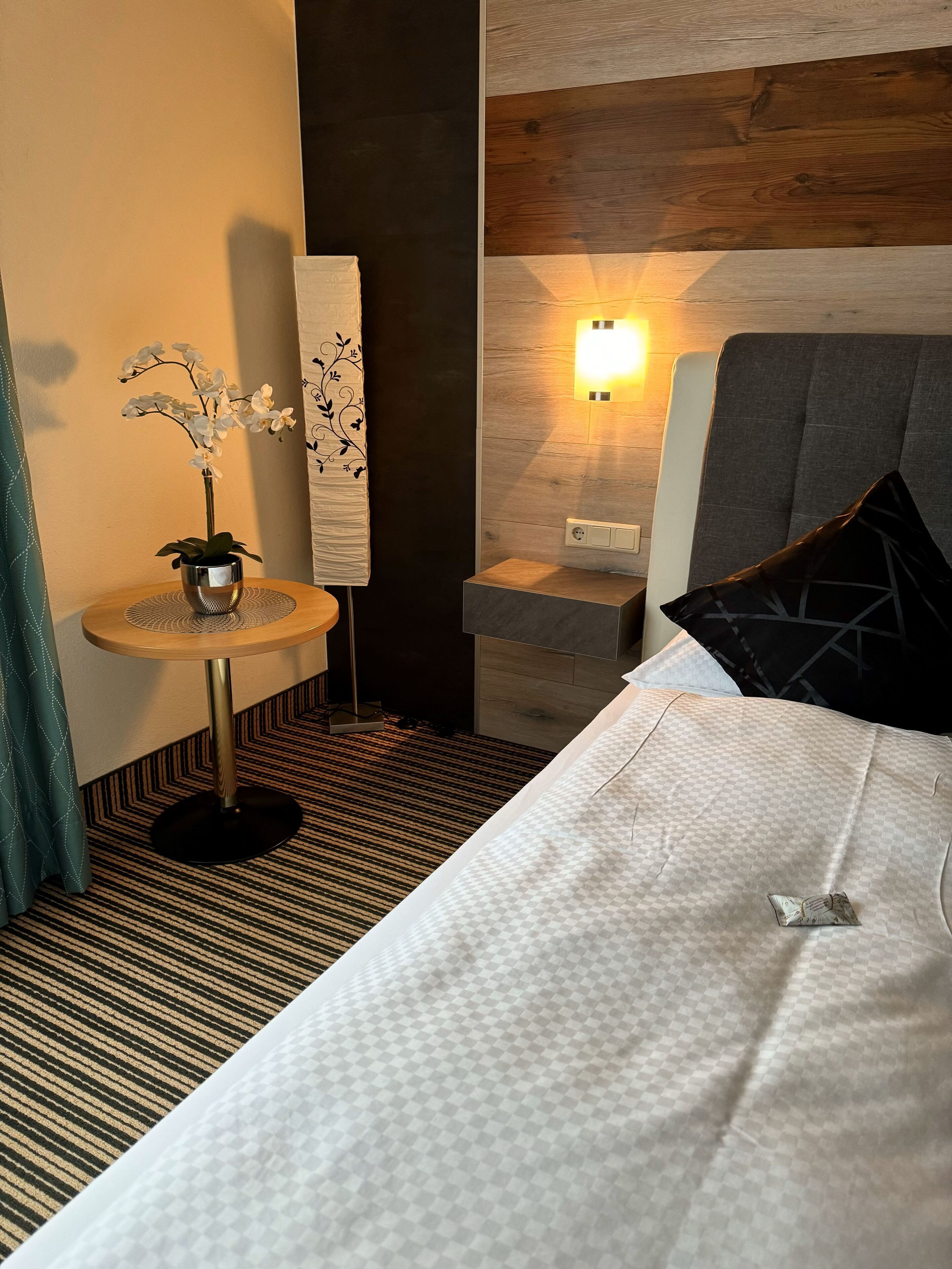 Ein Hotelzimmer mit einem Bett, einem kleinen runden Tisch mit einer Topfpflanze, einer Wandleuchte und einem gemusterten Teppich.