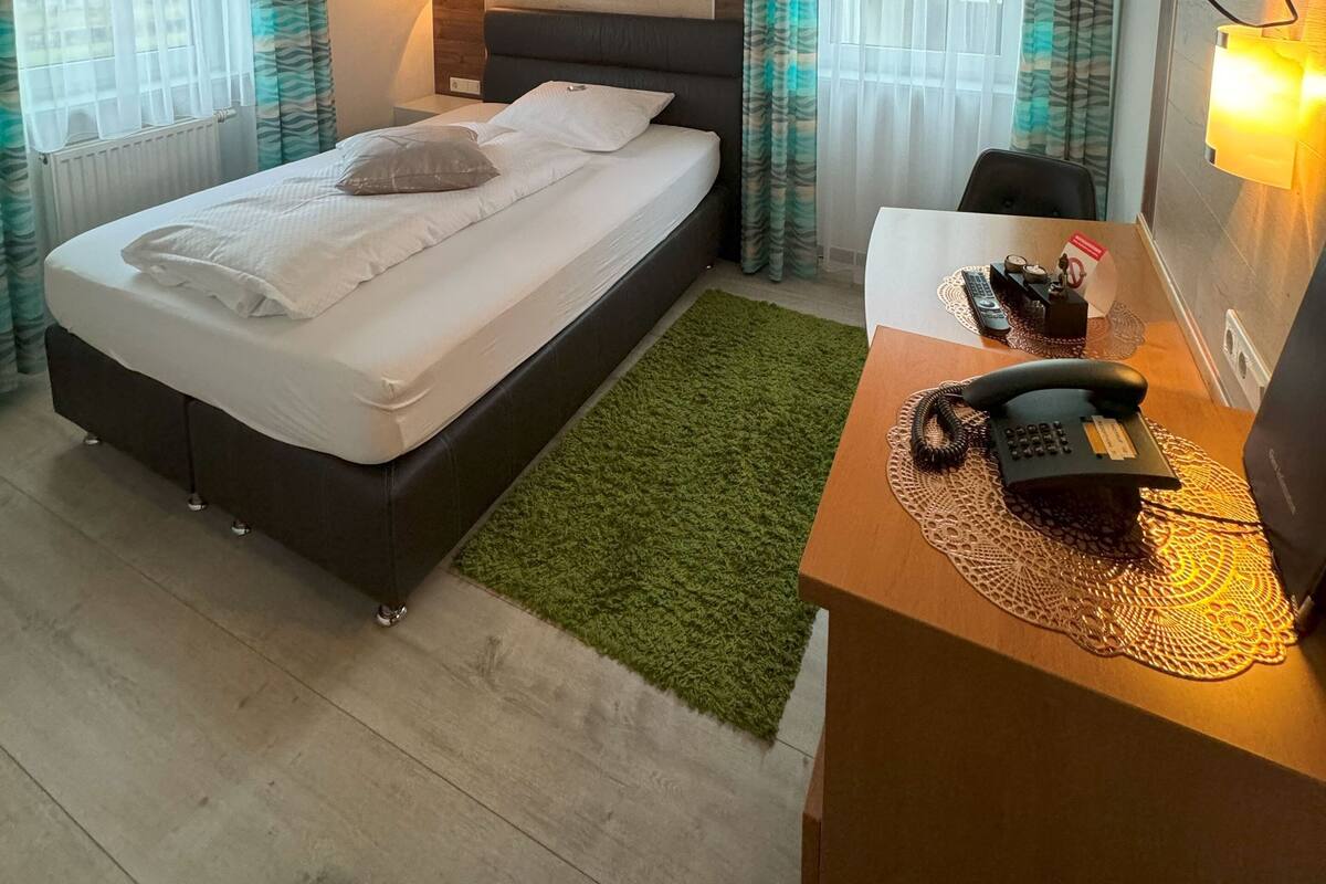 A hotel room with a bed, a desk with a telephone, a wall-mounted TV, and a painting of a forest.