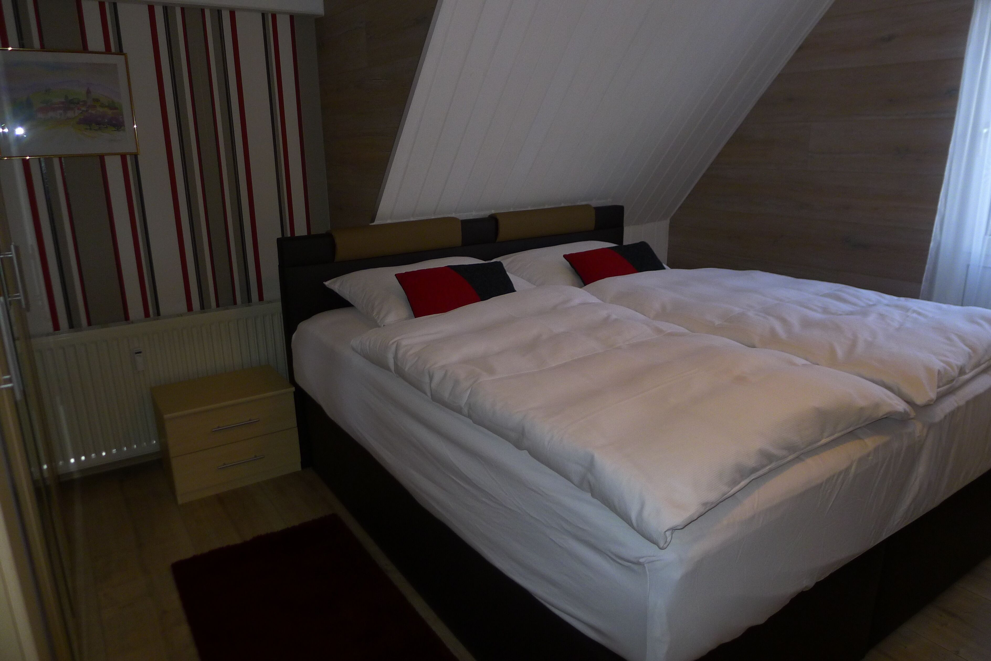 A neatly made bed with white bedding and red and black pillows, a wooden nightstand with drawers, and a radiator below.