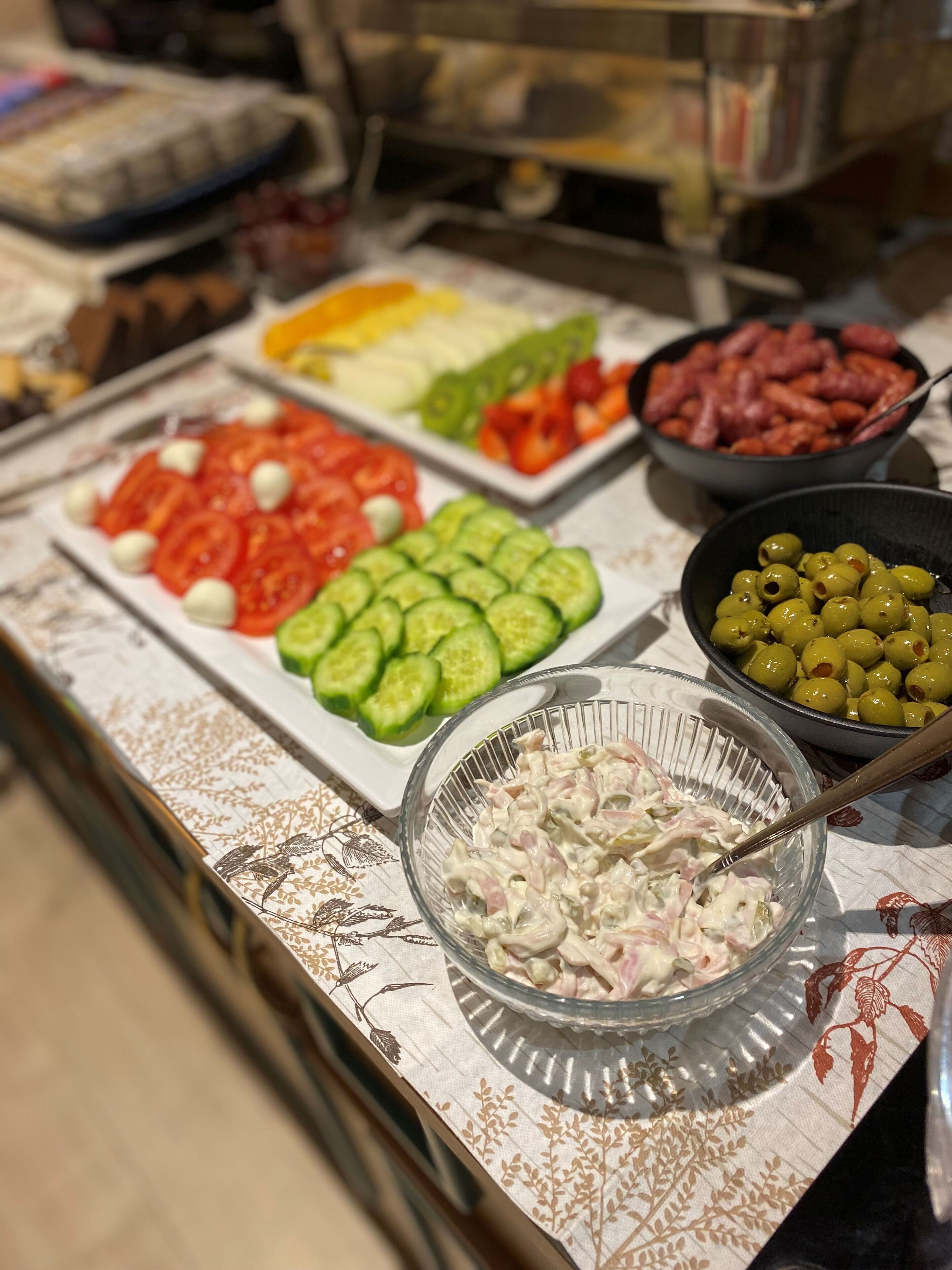 A buffet with various food items including sliced cucumbers, tomatoes, olives, and a bowl of salad.