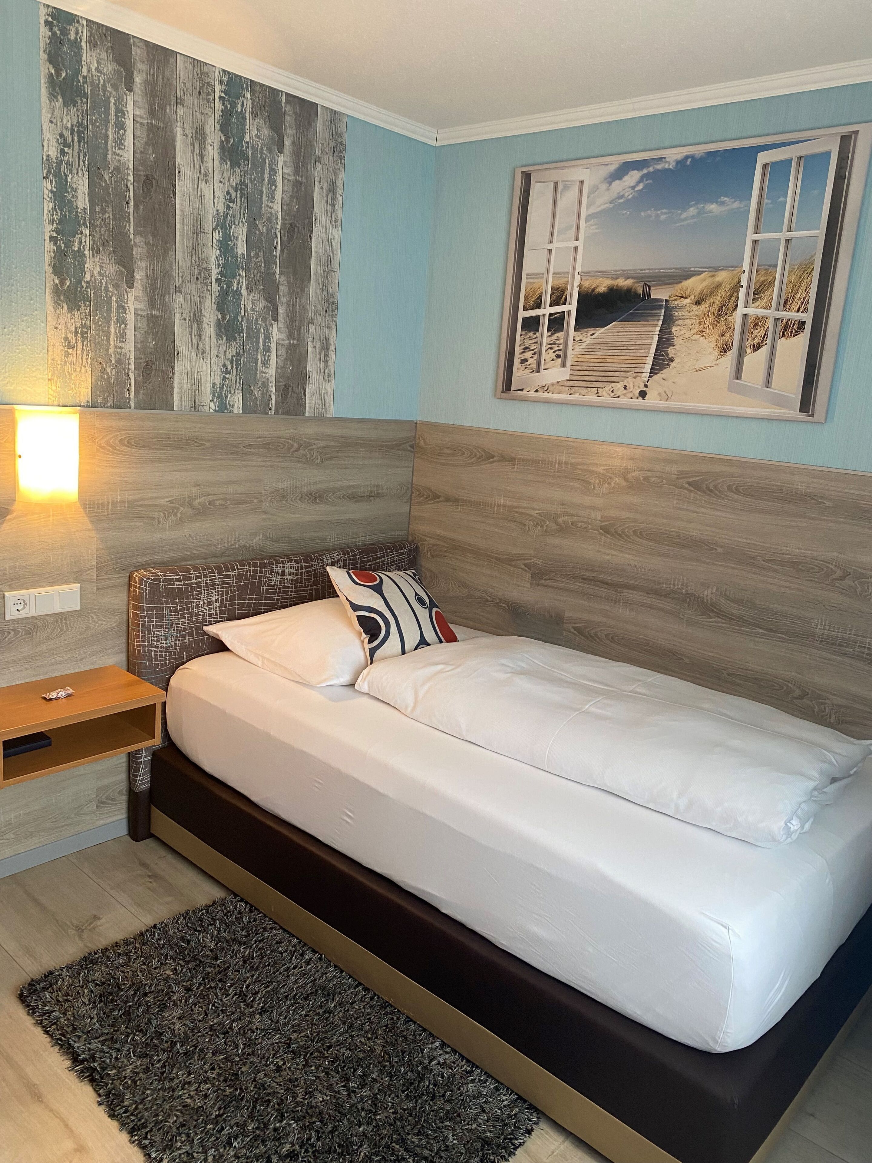 Een slaapkamer met een bed, nachtkastje, wandlamp en een foto met strandthema.