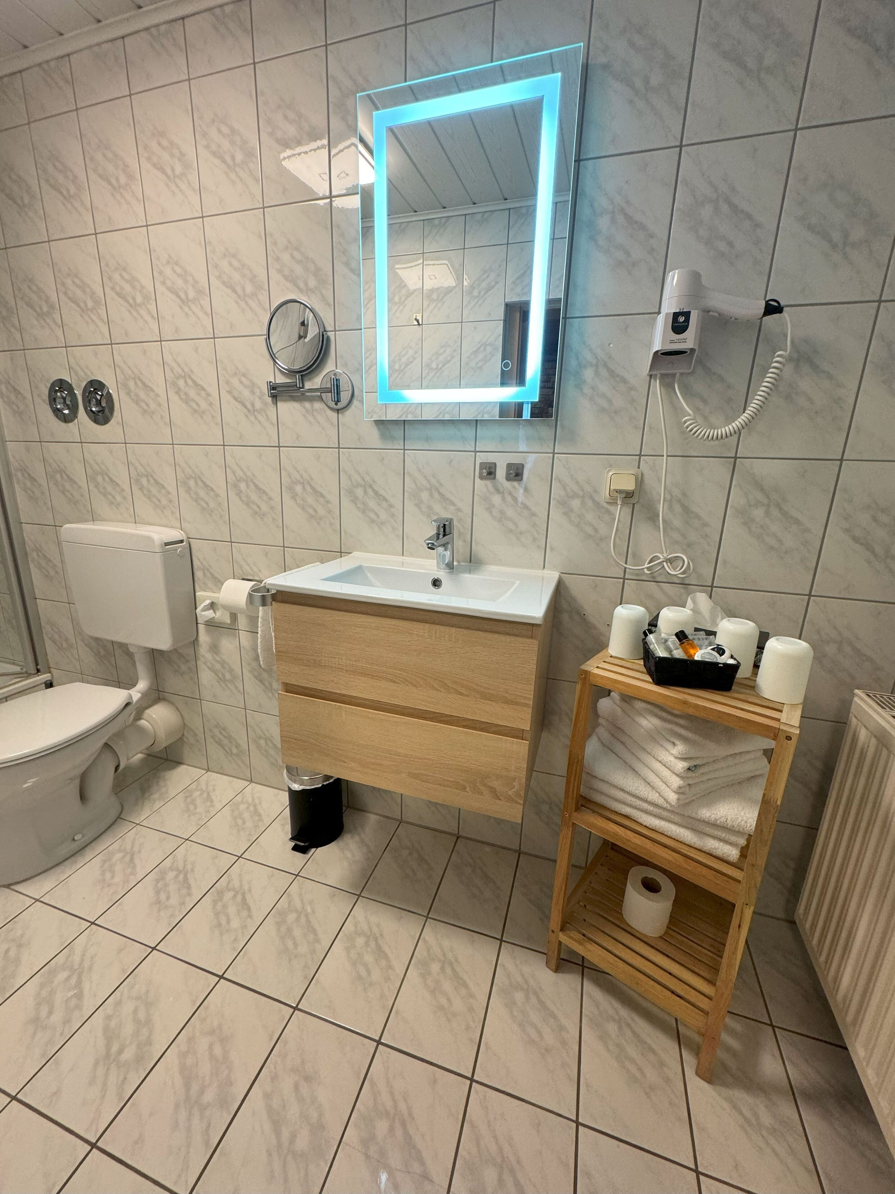 A modern bathroom with a white toilet, a sink with a wooden vanity, a mirror with a blue light, and a shelf with towels and toiletries.