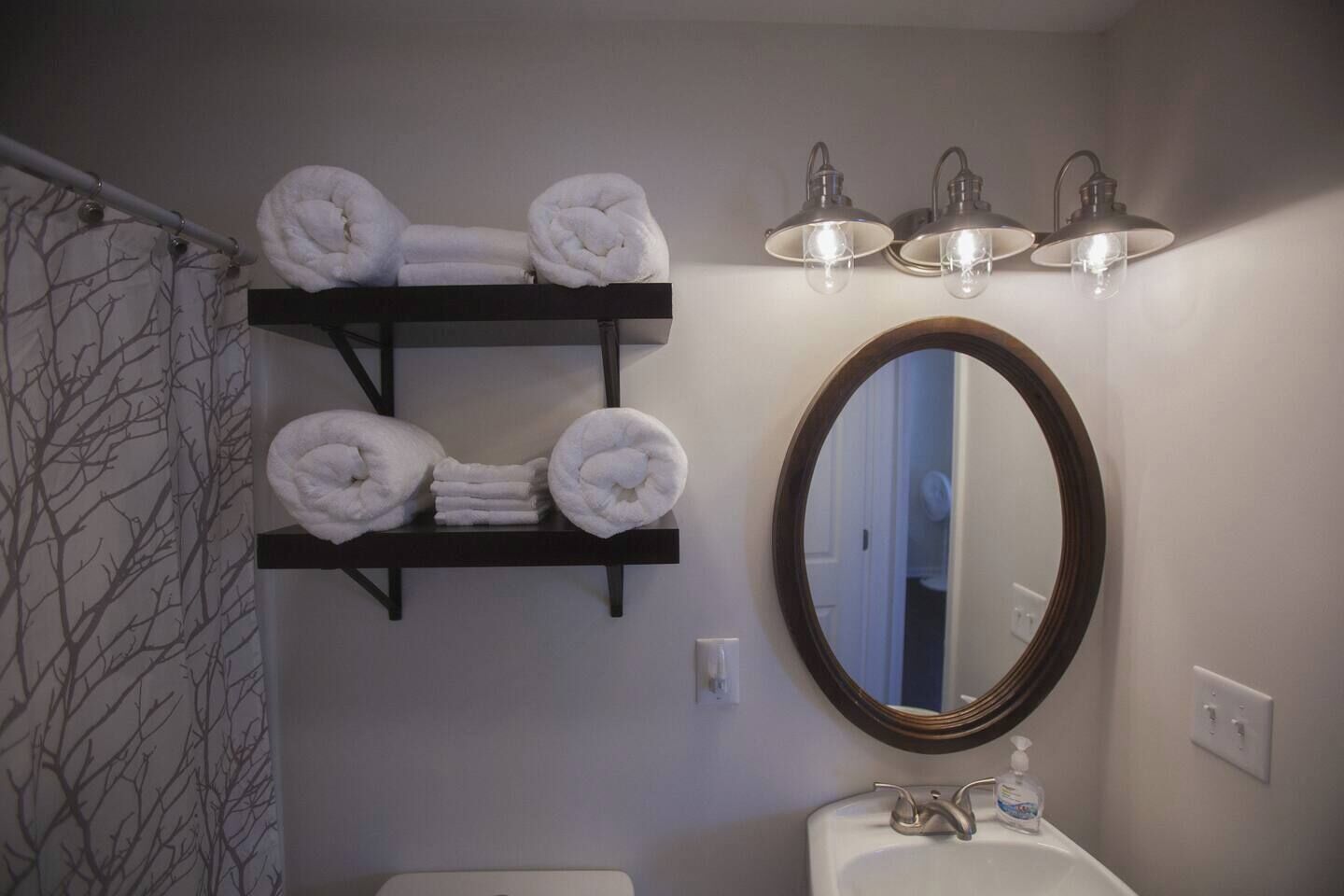 Une salle de bain avec un porte-serviettes où sont pliés les serviettes, un miroir rond et un lavabo équipé d’un distributeur de savon.