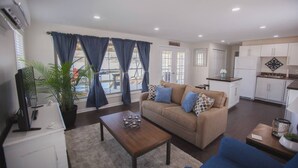 A modern living room with a beige sofa, a coffee table, and a TV. There is a kitchen area with white cabinets and a sink. - 2 Bedroom Lakefront Cabin with Hot Tub! (Brookport)