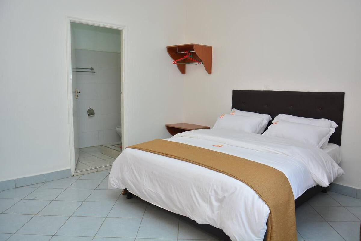 Una habitación de hotel con una cama, una mesita de noche, un estante de madera con un perchero rojo y una puerta que da a un baño.