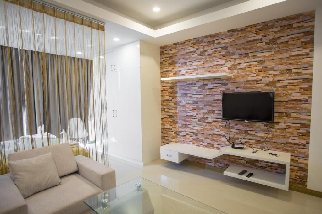 A modern living room with a sofa, a glass coffee table, a TV mounted on a stone-patterned wall, and a white entertainment unit with remote controls.