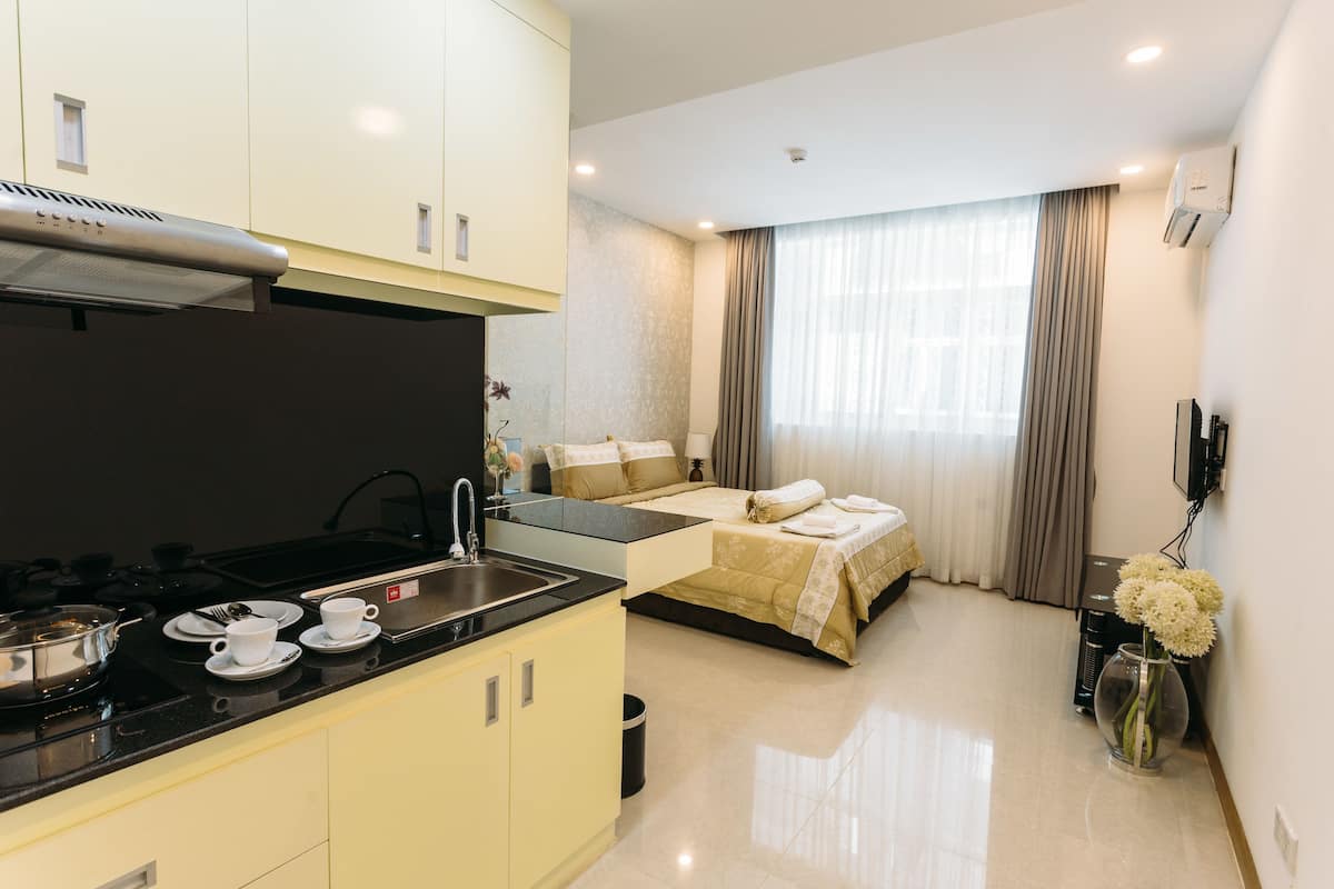 A modern hotel room with a kitchenette, a bed with a beige bedspread, a large window with curtains, and a vase with flowers on the floor.