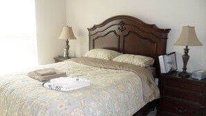 Un lit bien fait, recouvert d’une couvre-lit à motifs floraux, deux lampes de chevet, une table de chevet en bois avec un cadre contenant une photo, et une serviette pliée posée sur le lit.