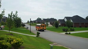 Un autobus scolaire jaune roule dans une rue de banlieue bordée de maisons et d’arbres.