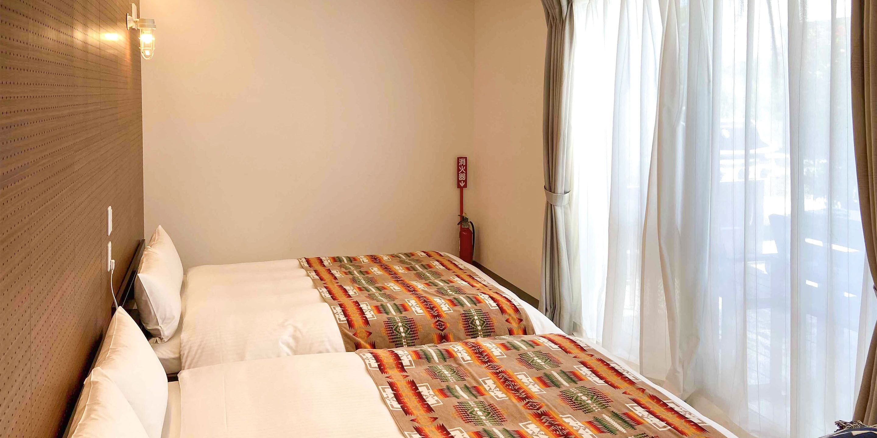 Ein Hotelzimmer mit zwei Betten, einem Holzkopfstück, einem Nachttisch, einer Lampe und einem Fenster mit durchscheinenden Vorhängen.