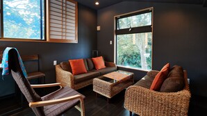 Ein Zimmer mit einer dunklen Wand, einem großen Fenster, einem braunen Sofa mit orangen Kissen, einem Korbstuhl und einem kleinen Tisch.