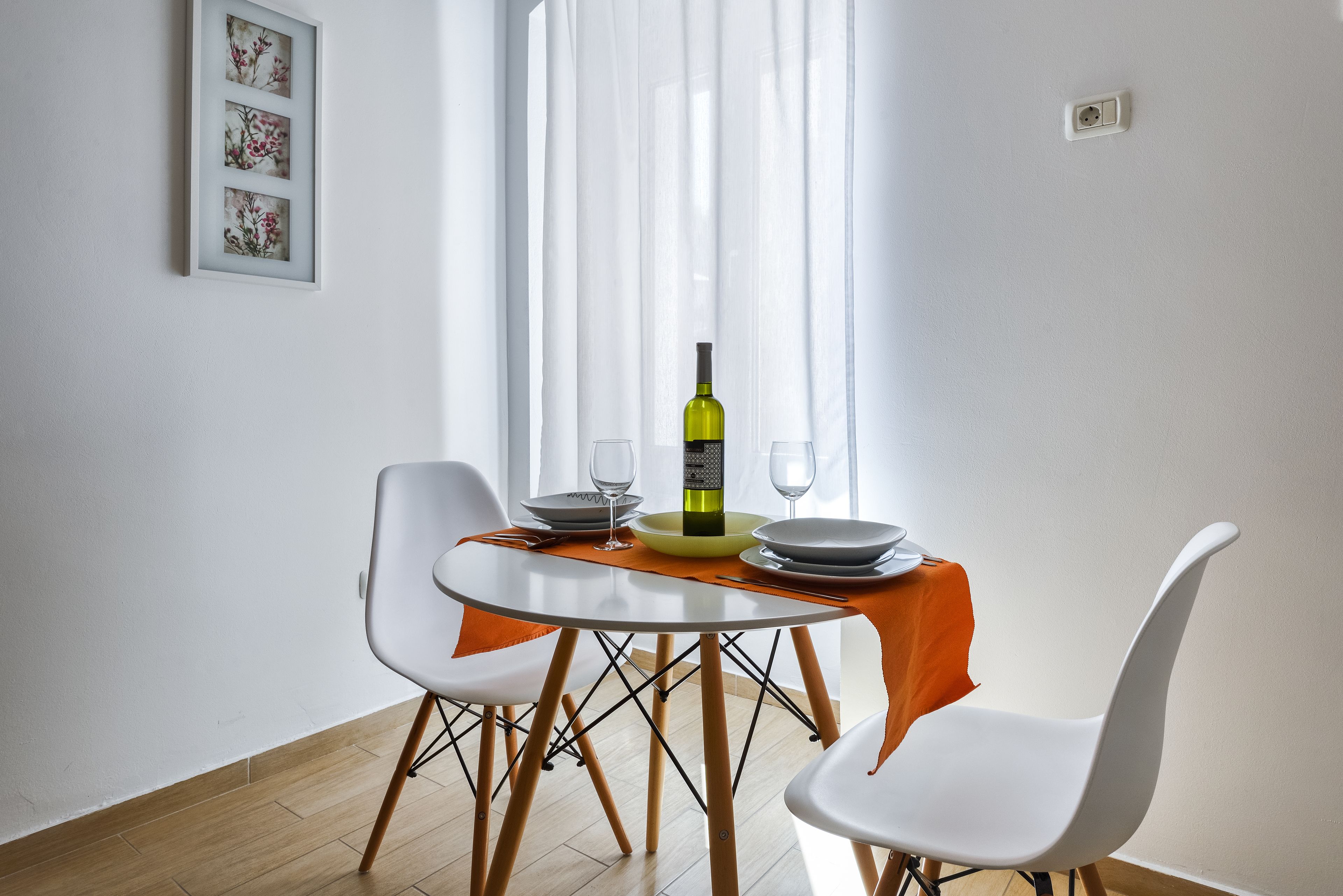 A dining area with a round table, white chairs, a bottle of wine, and a window with sheer curtains.