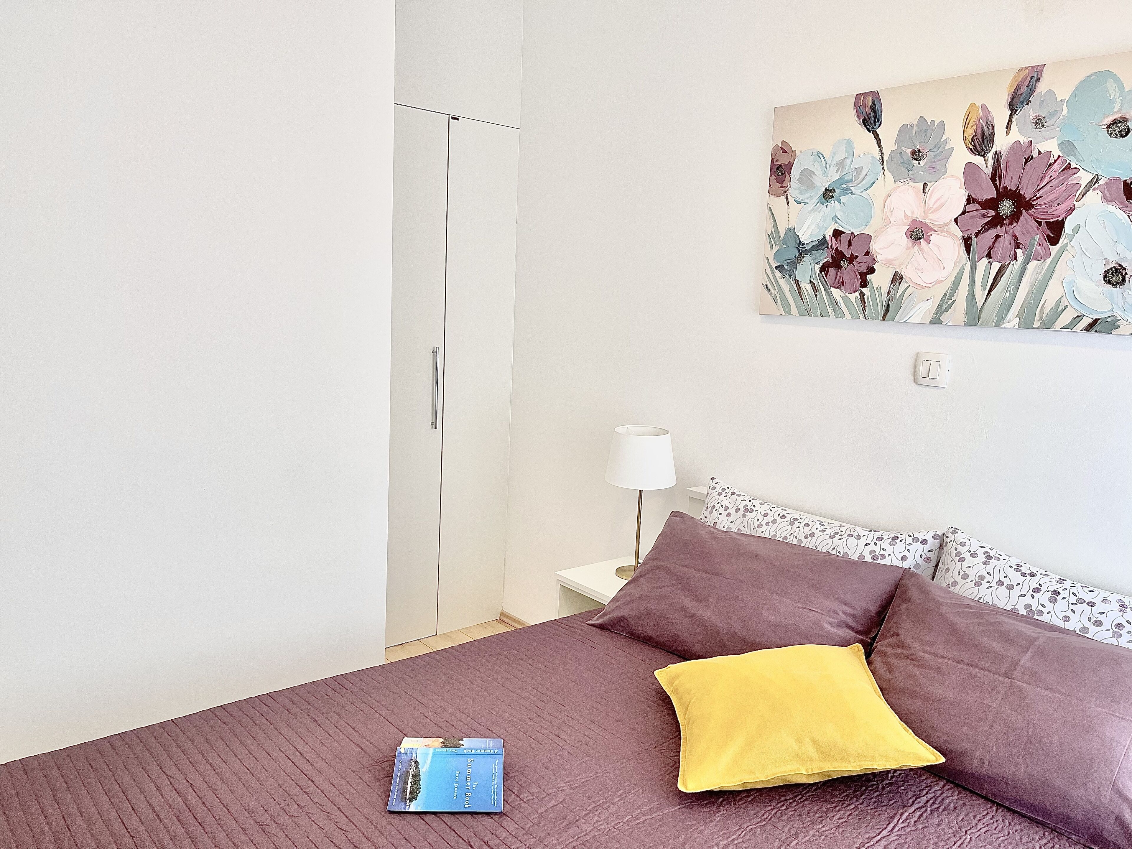 Una camera da letto con un letto, cuscini, un comodino, una lampada e un quadro appeso al muro.