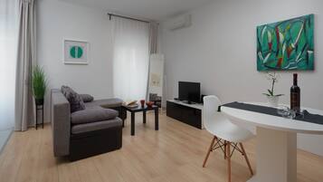 Una sala de estar moderna con un sofá gris, una mesita, una silla blanca, un televisor y un cuadro en la pared.