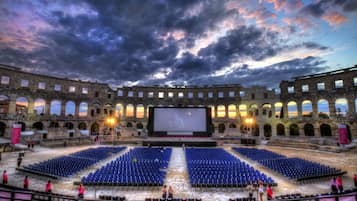 An outdoor amphitheater with a large screen, rows of blue seating, and stone architecture.