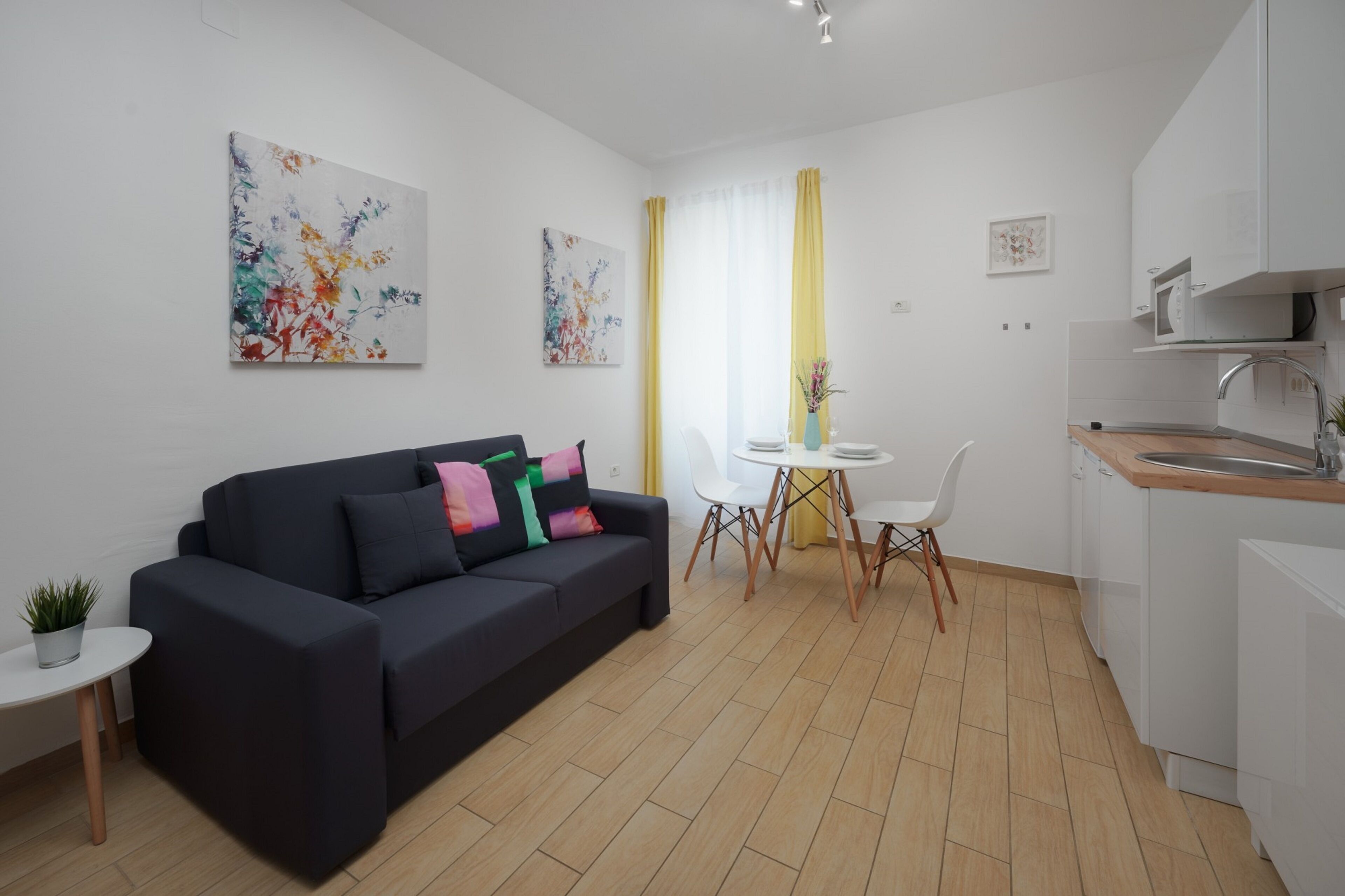 Una sala moderna con un sofá de color oscuro, una mesita redonda con una planta en maceta, y un comedor con una mesa y sillas blancas.