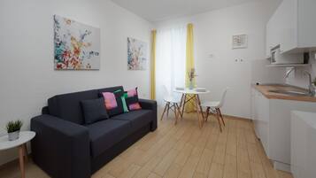 A modern living room with a dark-colored sofa, a small round table with a potted plant, and a dining area with a white table and chairs.