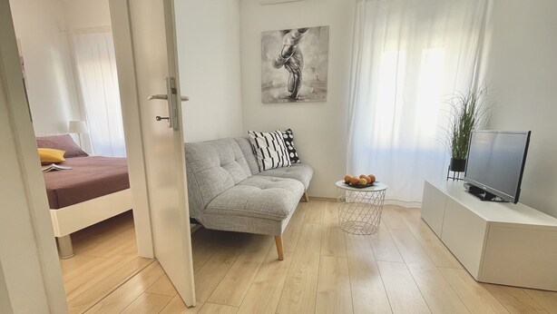 Un salon moderne avec un canapé gris, une petite table ronde sur laquelle sont disposés des fruits, un téléviseur à écran plat et un tableau accroché au mur.