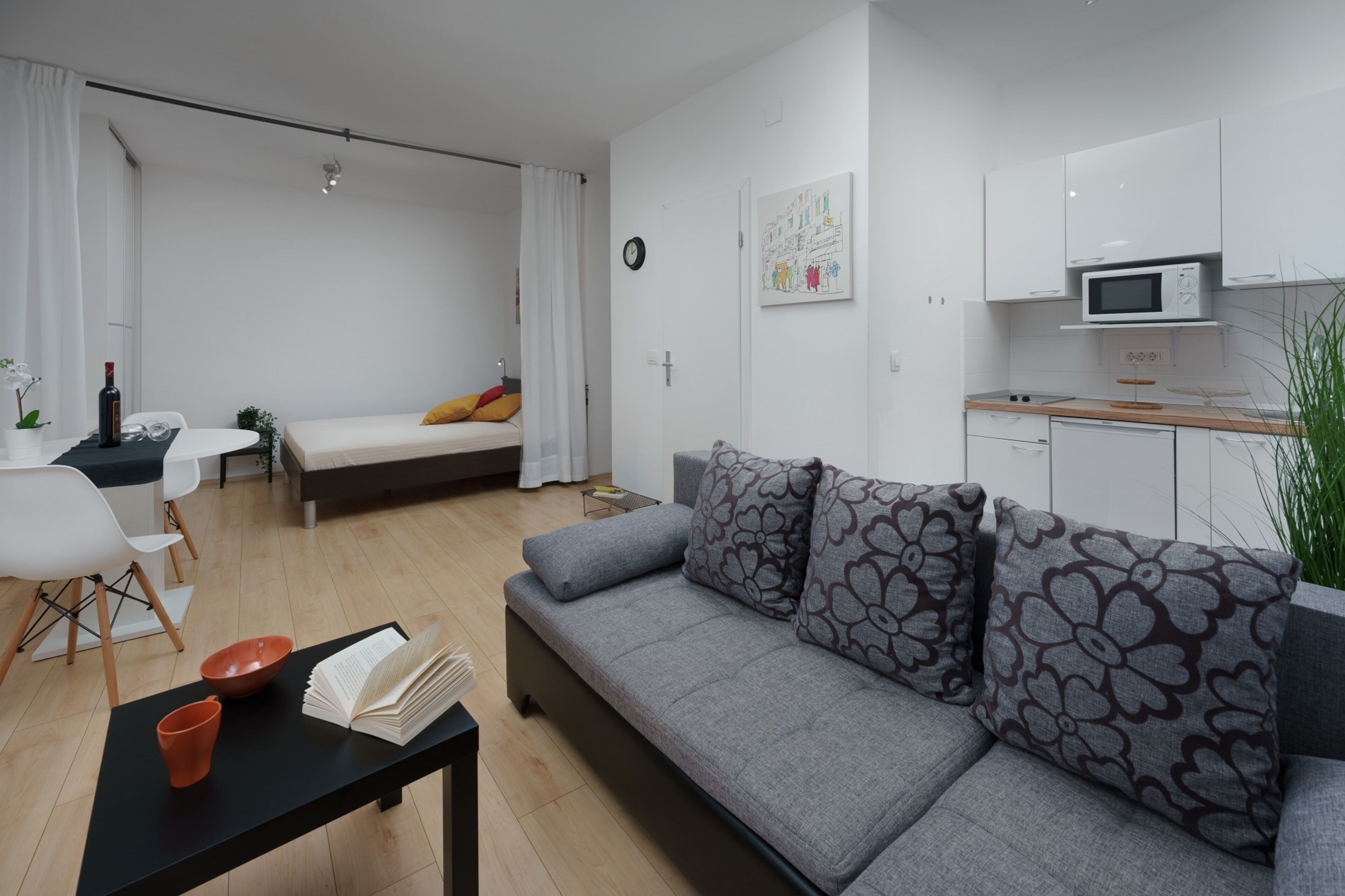 Un apartamento moderno con sofá gris, una mesa de comedor, una zona de cocina y una cama con cabecera blanca.