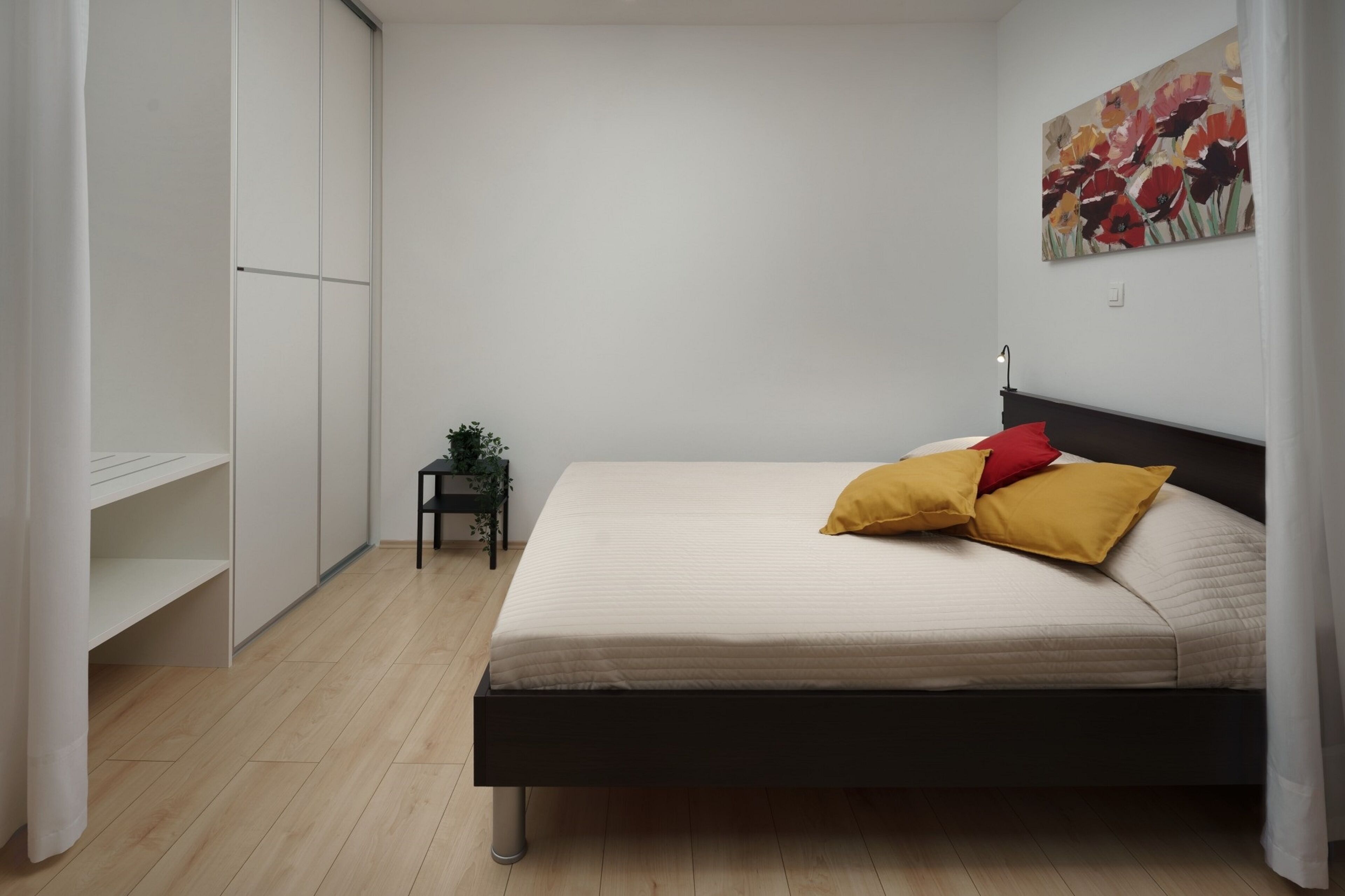 A bedroom with a bed, a nightstand, a small table with a plant, and a painting on the wall.