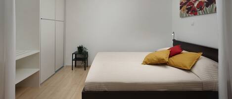 A bedroom with a bed, a nightstand, a small table with a plant, and a painting on the wall.