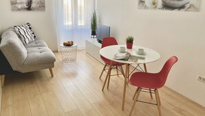 Un salon moderne avec un canapé gris, une table de salle à manger ronde entourée de chaises rouges, une petite table d’appoint sur laquelle se trouve un saladier de fruits, et deux tableaux accrochés aux murs.