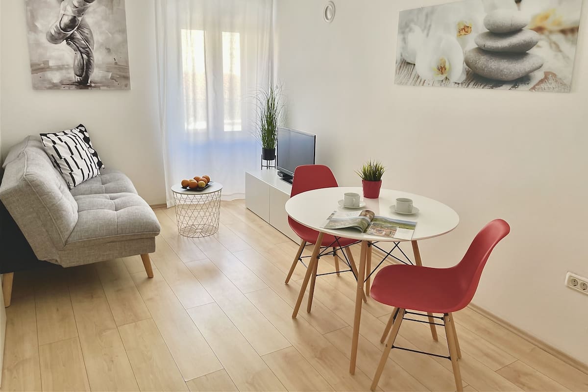 Un salon moderne avec un canapé gris, une table de salle à manger ronde entourée de chaises rouges, une petite table d’appoint sur laquelle se trouve un saladier de fruits, et deux tableaux accrochés aux murs.