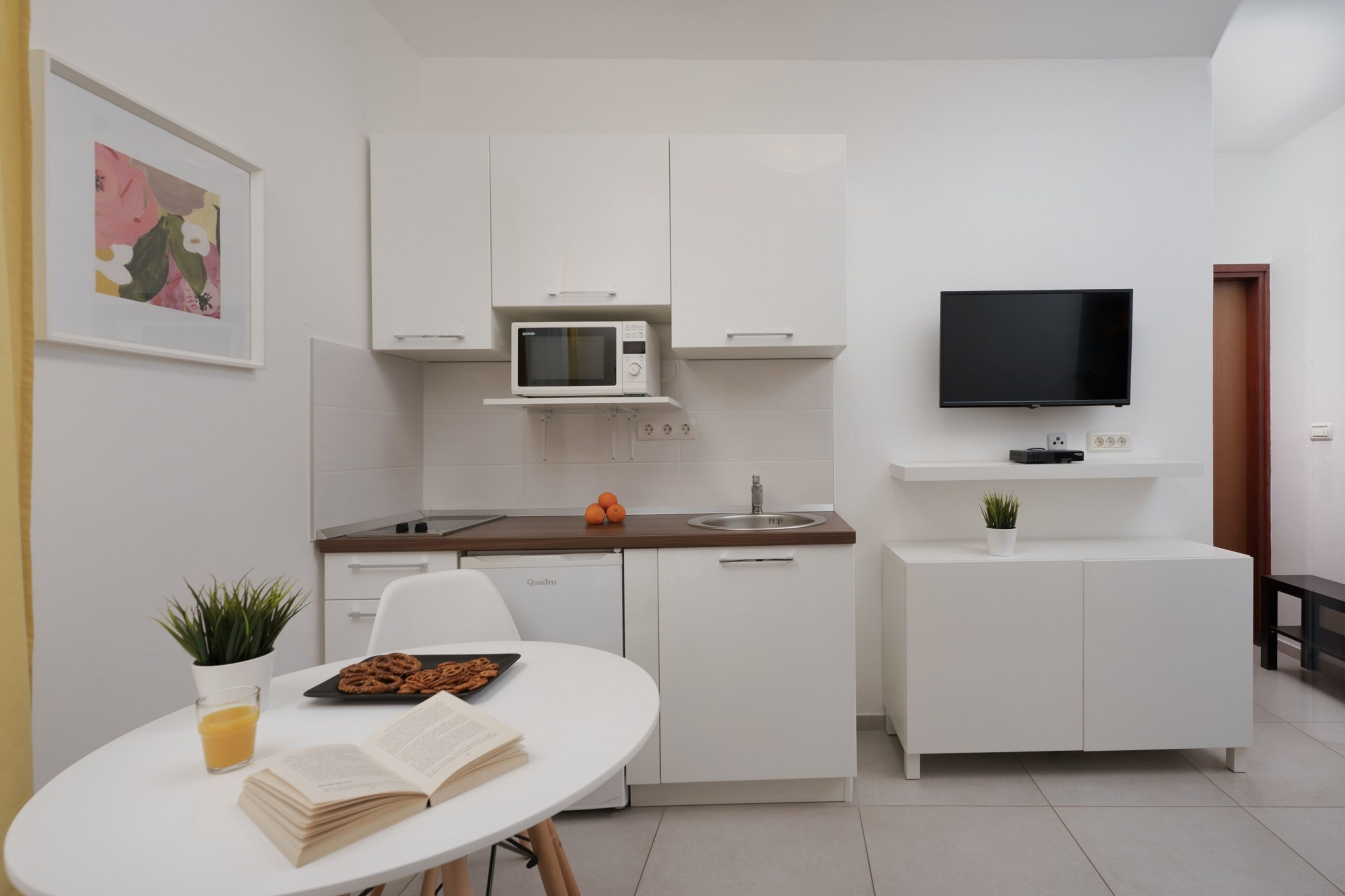 Een compacte keuken met witte keukenkastjes, een magnetron, een spoelbak en een televisie die boven de aanrecht is bevestigd.