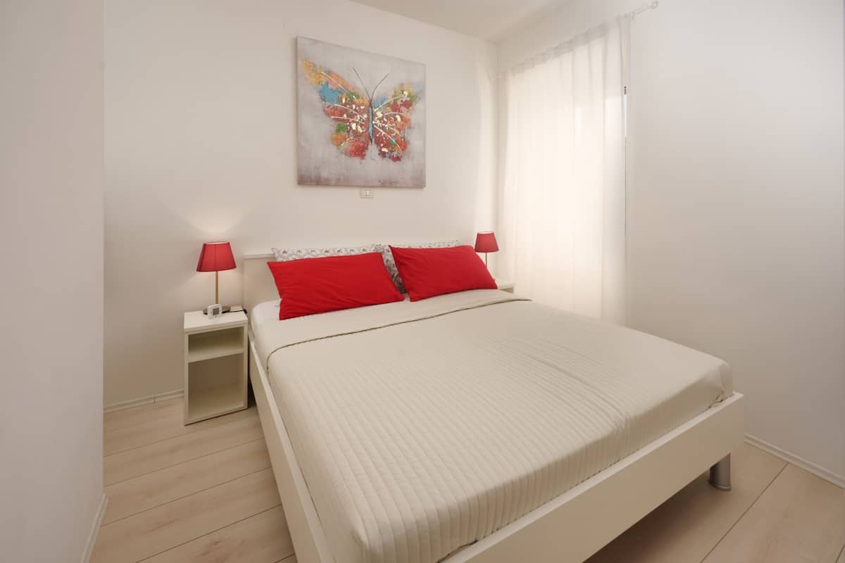 Un dormitorio con una cama, dos mesitas de noche, una lámpara roja en cada una, una pared blanca y un cuadro de una mariposa colorida.