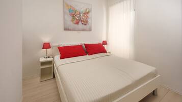 Un dormitorio con una cama, dos mesitas de noche, una lámpara roja en cada una, una pared blanca y un cuadro de una mariposa colorida.