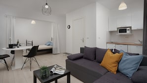 Una sala de estar moderna con un sofá gris, una mesa de comedor con sillas, una pequeña cocina y un dormitorio independiente con cama y cortinas.
